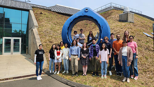 Large group of people standing in front of a large, circular, blue sculpture and a door.